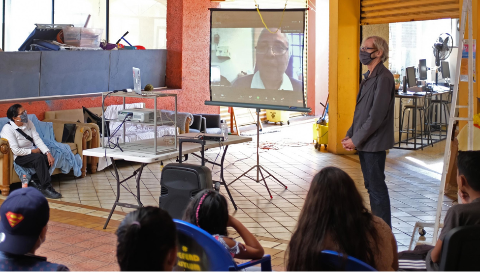 A group is listening to a presentation of a person talking on a projector in a Zoom screen. The presenter is wearing a mask and standing next to the projected screen.
