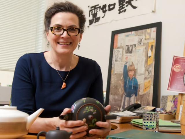 Professor Katharine Burnett sits at her desk and is holding a tea pot.