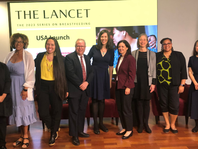 Nine individuals standing in a horizontal line smiling for a group photo. Each individual is wearing professional attire. They pose in front of a screen that reads “THE LANCET The 2023 Series on Breastfeeding USA Launch”