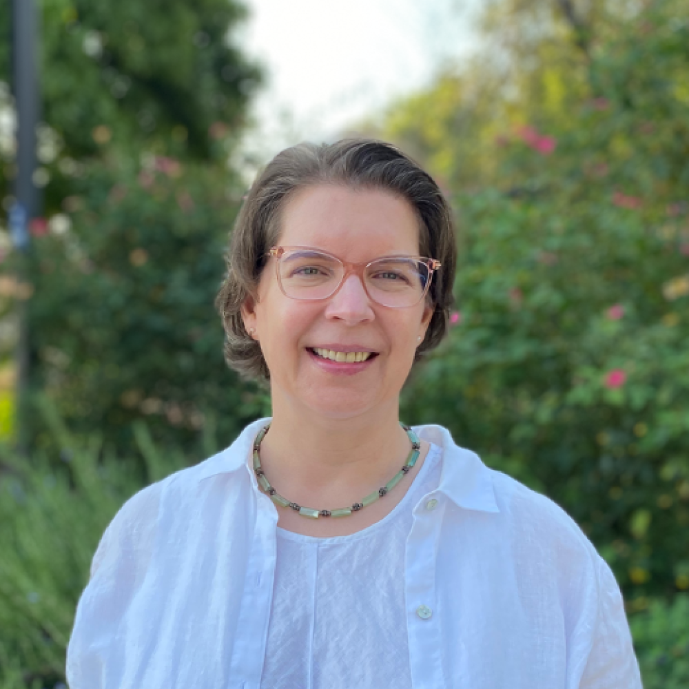 Becky Oskin, woman with glasses and a necklace wears a button down shirt. She smiles directly into the camera outside in front of bushes of flowers and trees. 