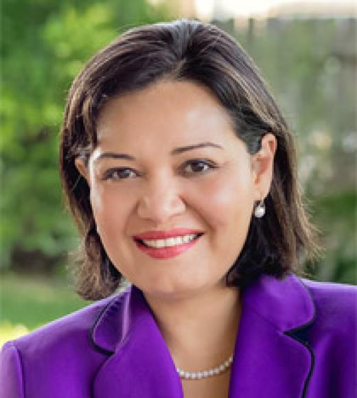 Woman in a bright purple blazer smiles directly into the camera in front of a blurry nature background
