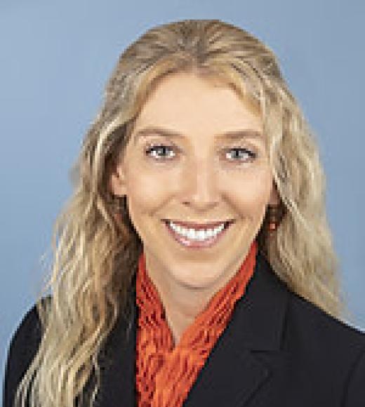 Woman wearing a blazer and collared shirt smiles directly into the camera in front of a plain background
