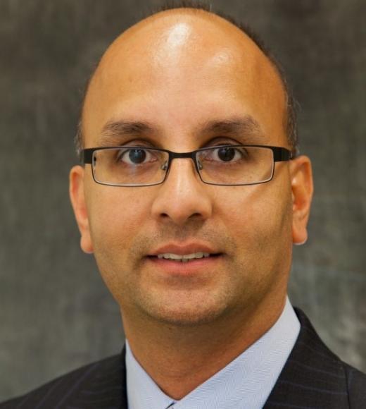 Man wearing glasses and a suit looks directly into the camera closeup in front of a plain background