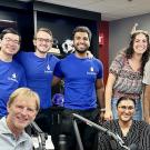 Mark Haney sits in a radio studio surrounded by several Square Solutions founders, students and entrepreneurs Henry Yu, Dillon Hill, Akshaj Raghavi, Sophie Bloyd, Harjn Bains and Deesha Patel.