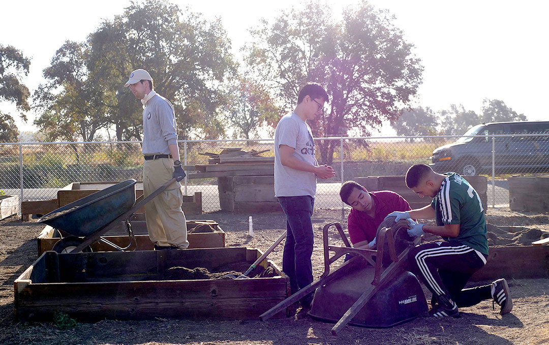 one man in a hat carries a wheelbarrow while three individuals look at a broken wheelbarrel