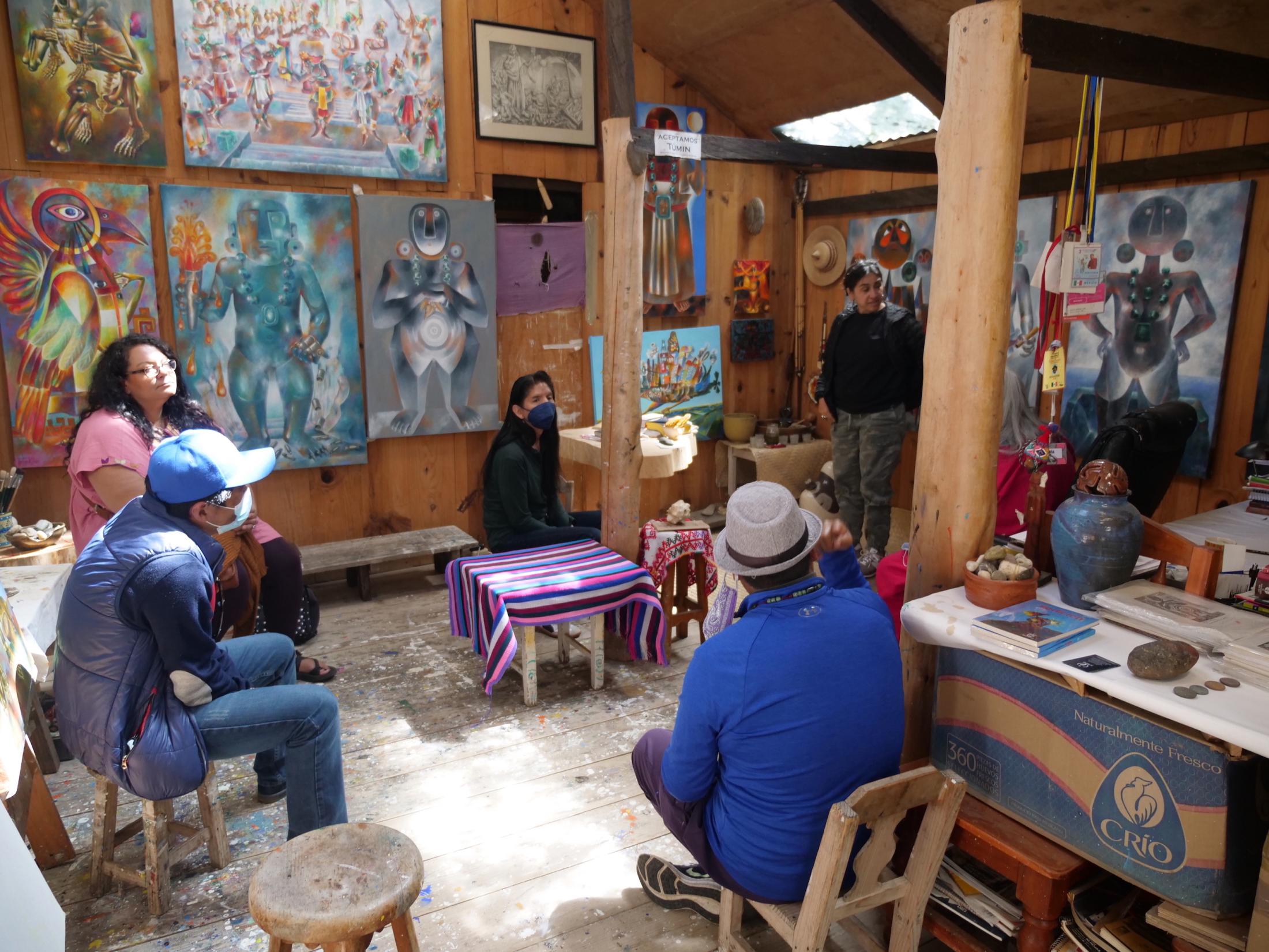 Group of individuals gather in a circle inside a studio full of colorful paintings by Antun Kojtom.