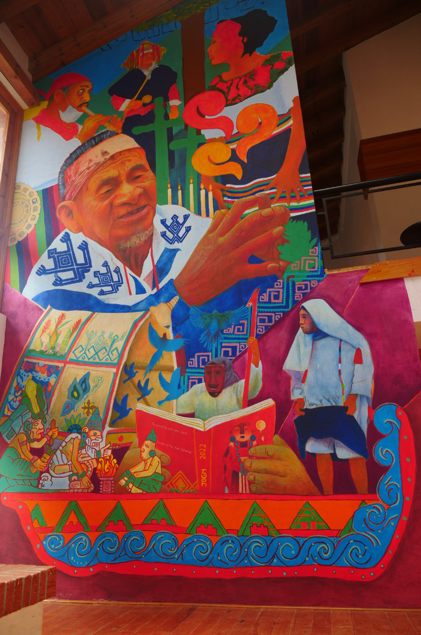 Colorful mural of people together in a collage. Three individuals at the left bottom are gathered to tell stories. There is a book in the center that is open. An elderly man at the upper half of the mural puts his hands together. Three individuals above the elderly man play music together. 