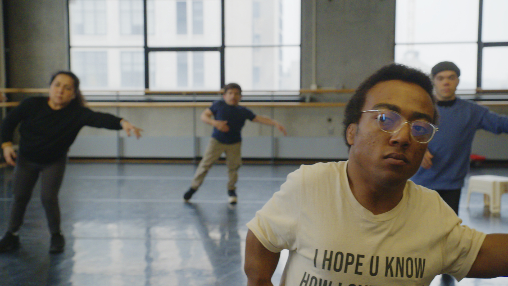 Inside a dance studio, four individuals are dancing with their arm reaching out as they learn the choreography.