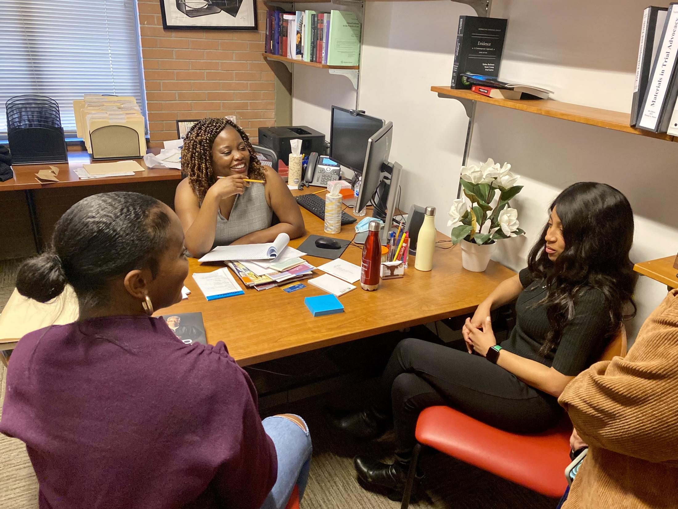 Professor Irene Joe sits at her desk to talk to two women in her office. She holds a pencil to take notes. 