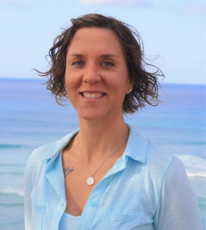 Woman with short hair wearing a light blue button up shirt and necklace smiles directly into the camera in front of a blurry ocean background