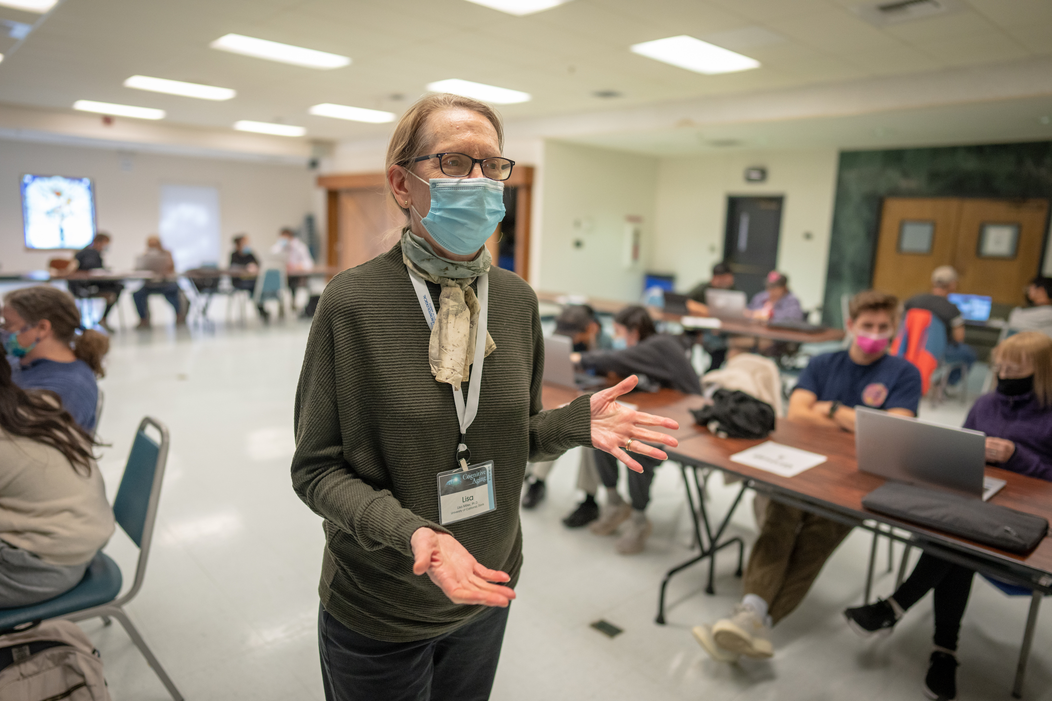 Faculty woman, Professor Lisa Miller, has blond hair tied in a ponytail. She is wearing glasses and a mask. She is in a classroom and is talking with both her hands out. Students are seated with computer in the background.