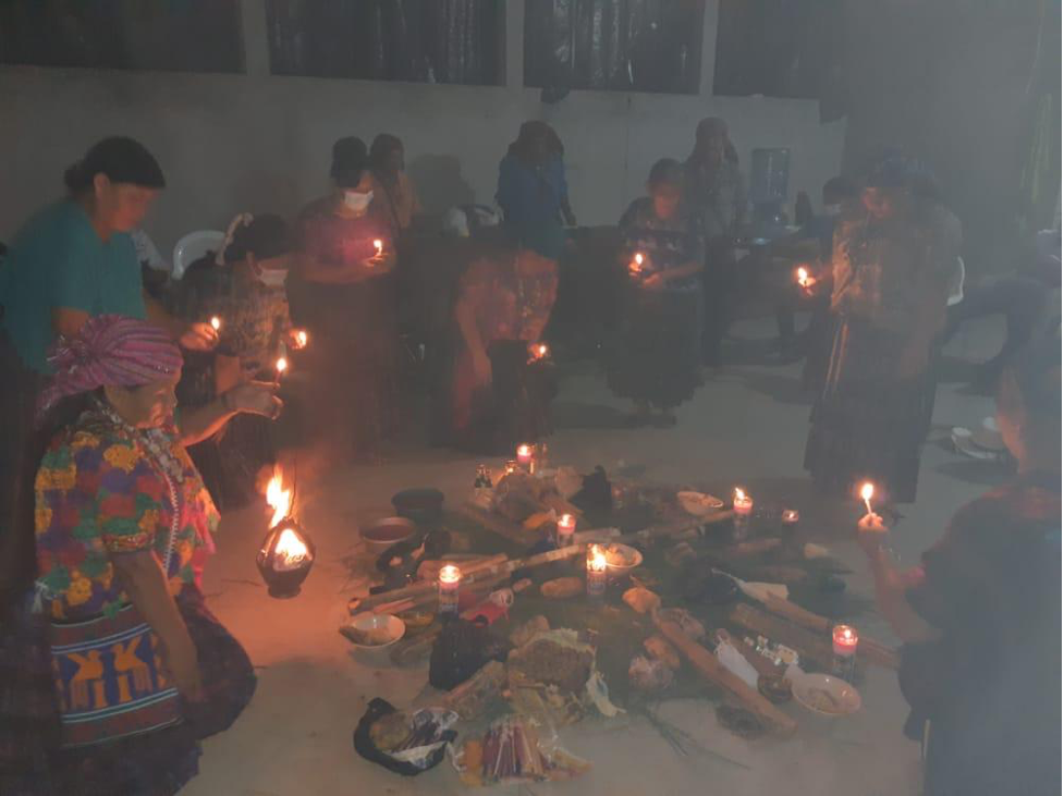 Group of people stand in a circle outside in the dark around items on the group. Most are holding an incense. 