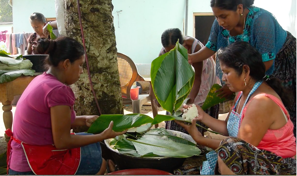 Three women sit together holding large leaves with rice