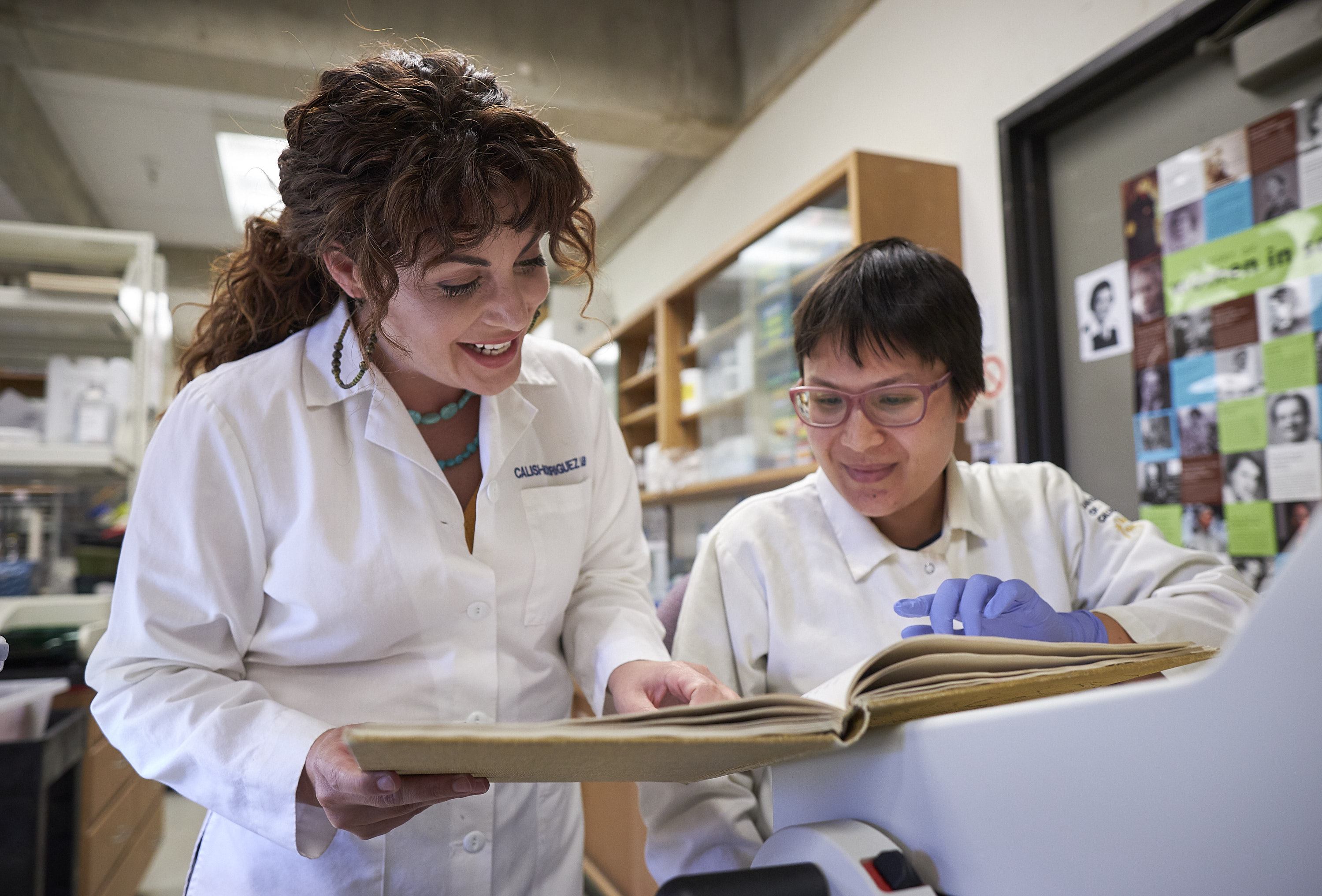 Two women in a lab wearing white lab coats look at a book together