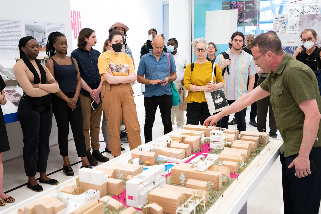 A group stands behind a large architectural site model while a man points at the model.