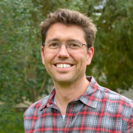 Man wearing glasses and a flannel plaid button up shirt smiles directly into the camera in front of a blurry nature background