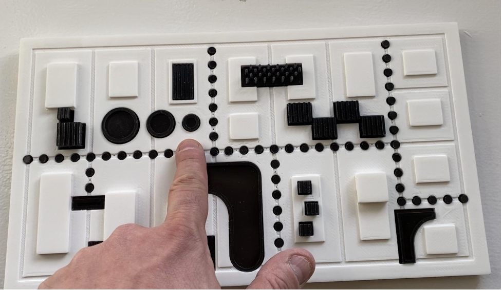 A finger points to a close up of a tactile model that includes various shapes on top of a flat surface