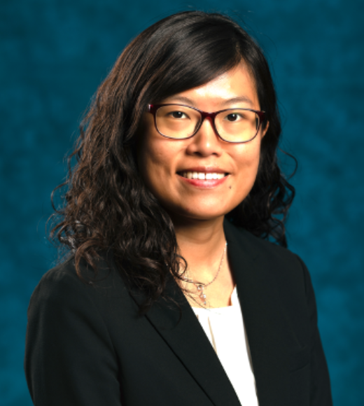 Close up of a woman wearing glasses and a blazer. She smiles in front of a plain background.