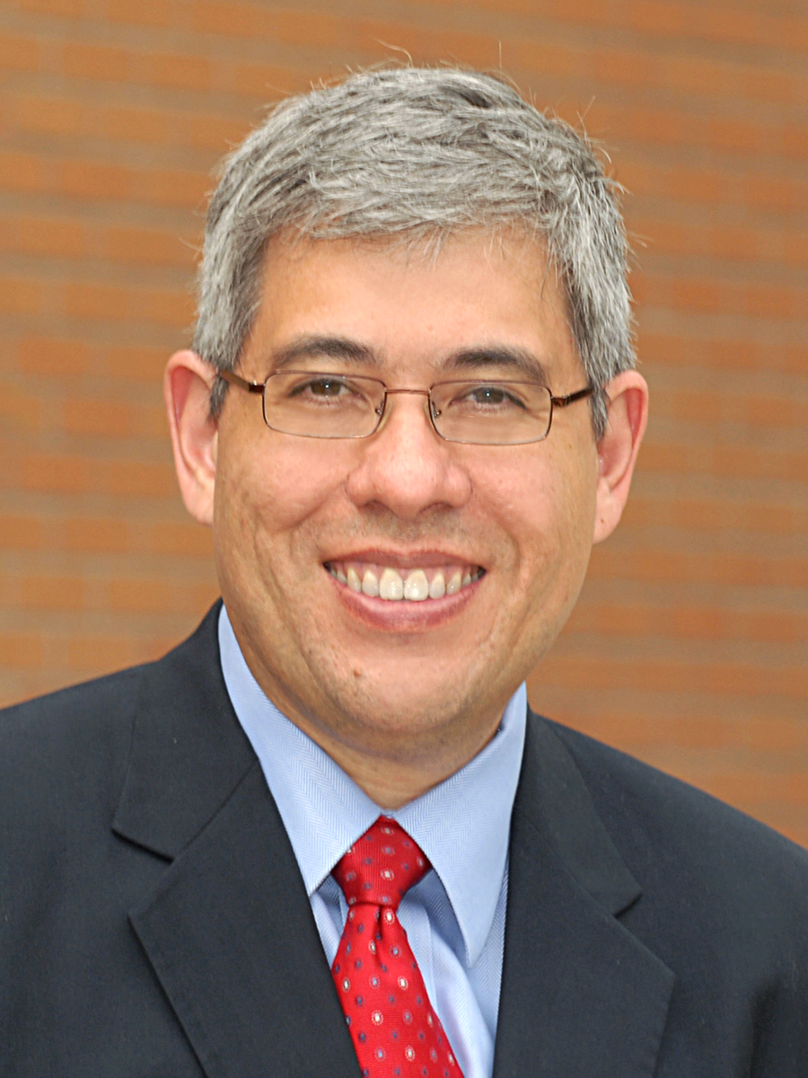 Gabriel “Jack” Chin wearing a suit and red tie smiling in front of a brown background