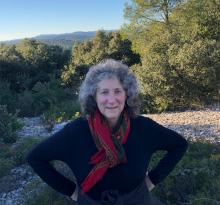 Diane Ullman smiling in front of a nature scenery. She wears a red scarf and a black long-sleeves shirt.