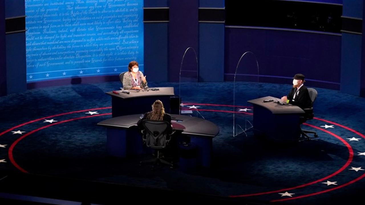 a debate stage with three people sitting far apart wearing masks