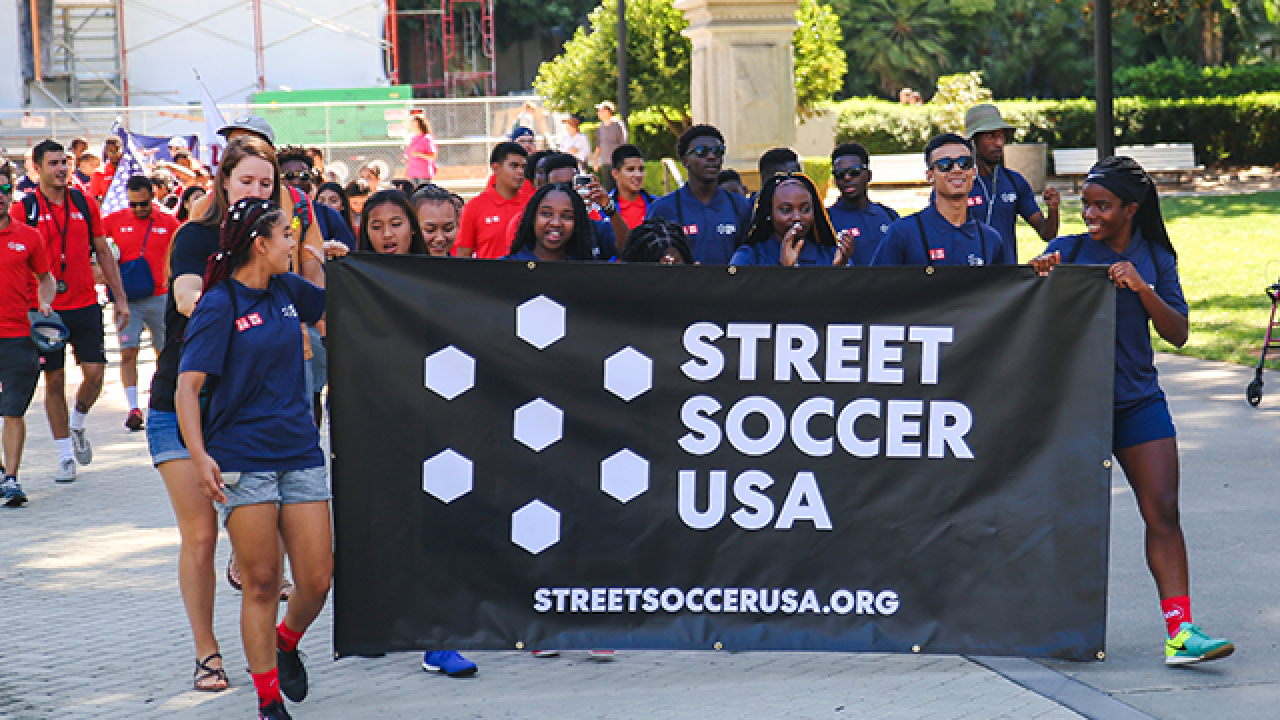 Individuals wearing blue polo shirts smiling while holding a Street Soccer USA sign with the words "Street Soccer USA STREETSOCCERUSA.ORG"