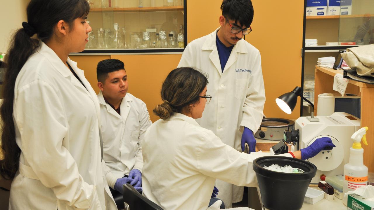 Four students in a white lab coat testing a lab equipment