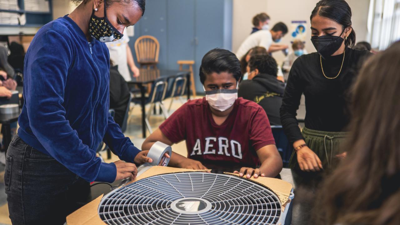 three high school students in masks looking at a large fan
