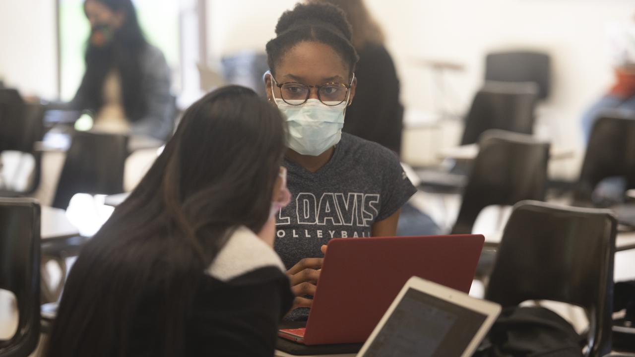 two students are talking to one another in front of their laptops