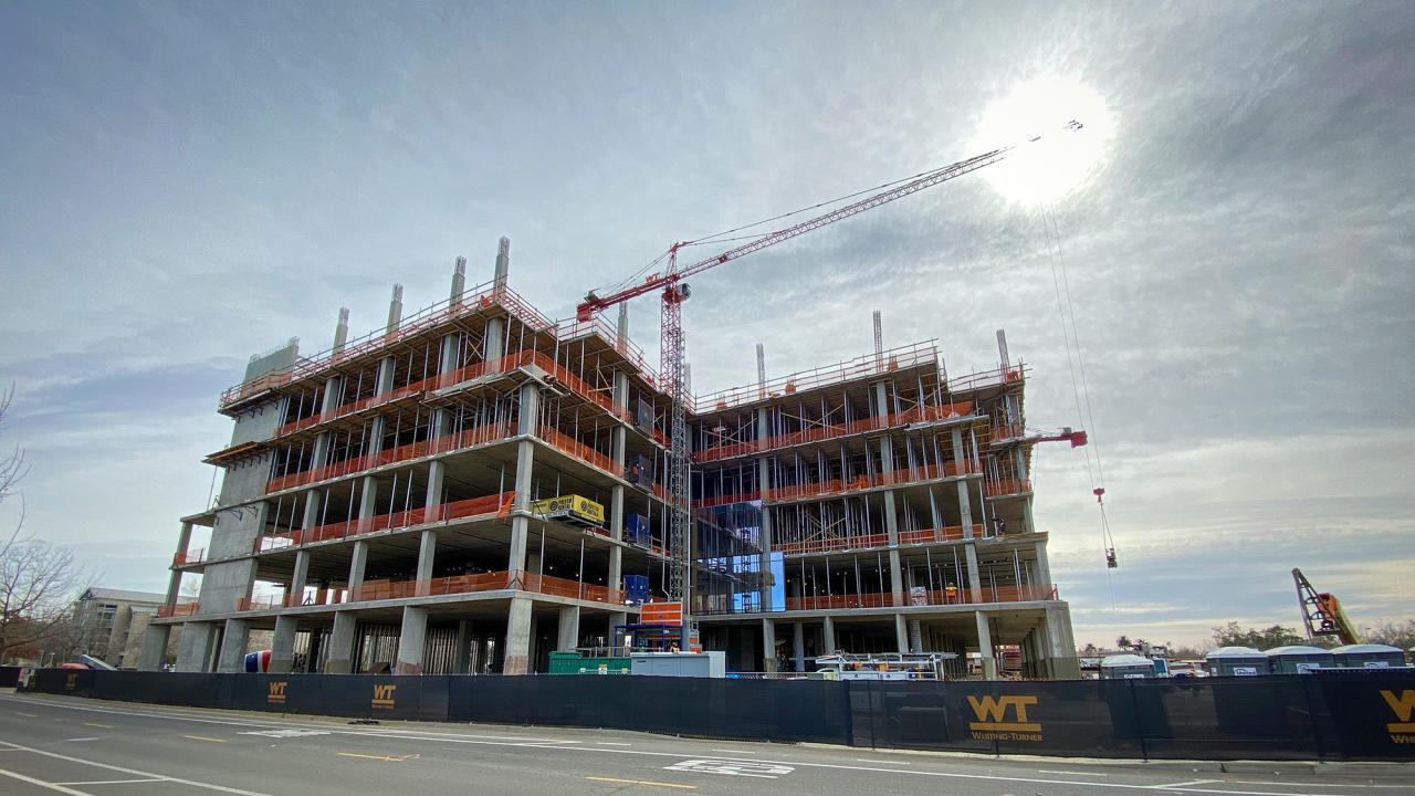 alt="Aggie Square, a UC Davis building on the Sacramento campus, is undergoing construction. The building's framing comprised of structural beams peaks into the partially cloudy sky."