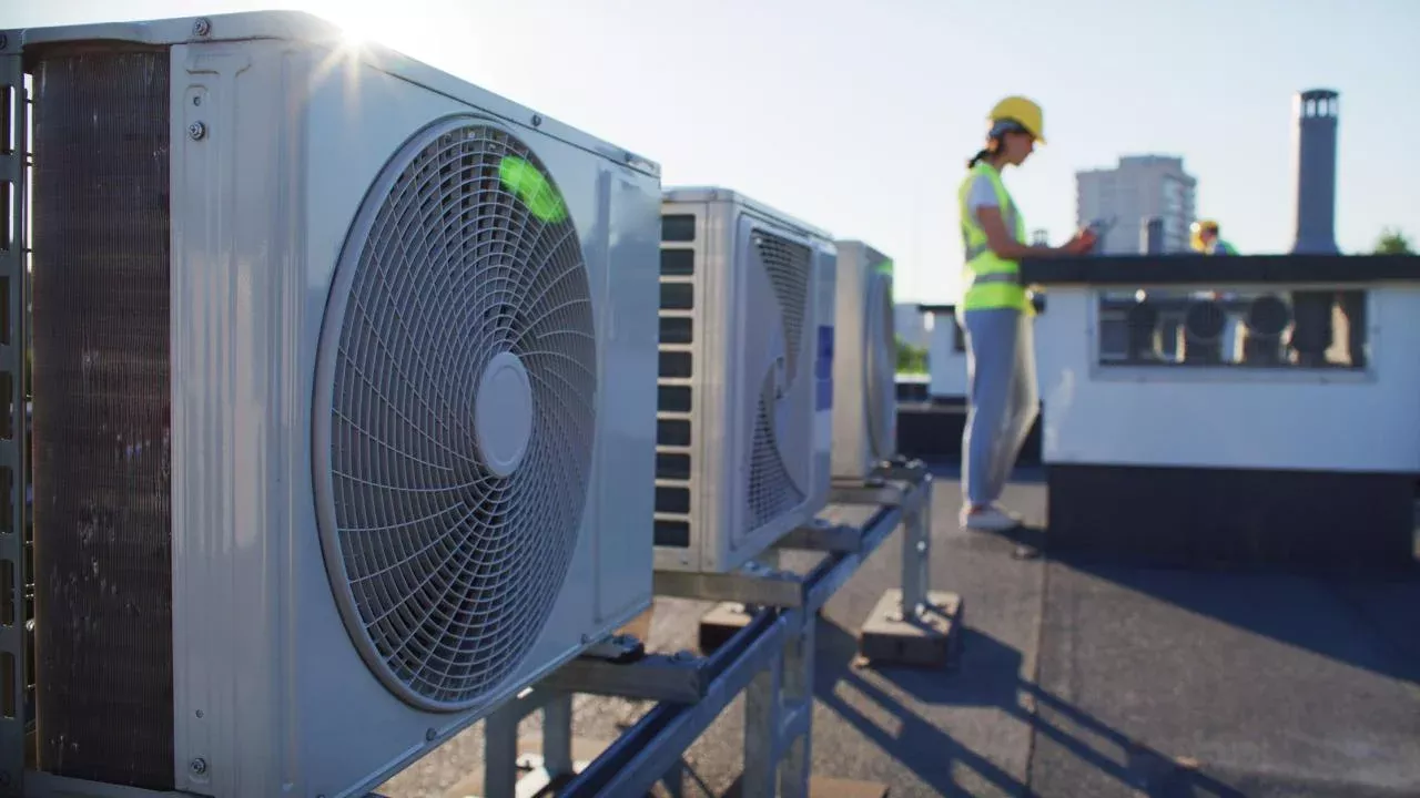 Oblique view of air conditioner units on a rooftop. A technician wearing a high visibility vest and hard hat is in the background.