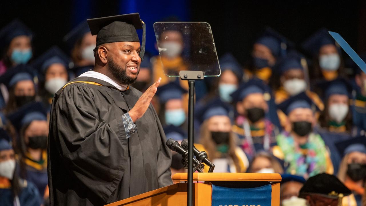 A person wearing a graduation cap and gown speaks at a podium with a UC Davis banner. They are holding up one hand, appearing to gesture or take an oath, while addressing the audience through a microphone. In the background, other graduates in caps and gowns sit, with some wearing face masks.