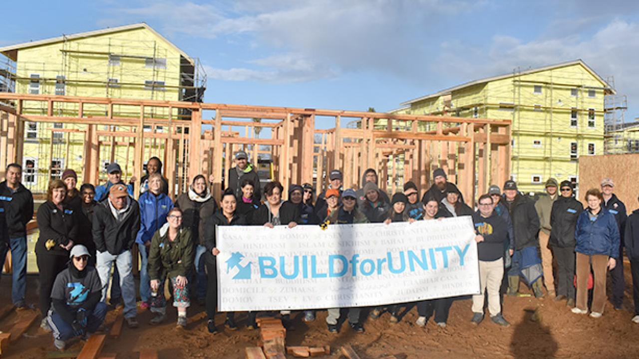 A group of about 40 people, most wearing coats and many wearing hats, stand together at a homebuilding site, behind a large white banner with the text "Build for Unity"
