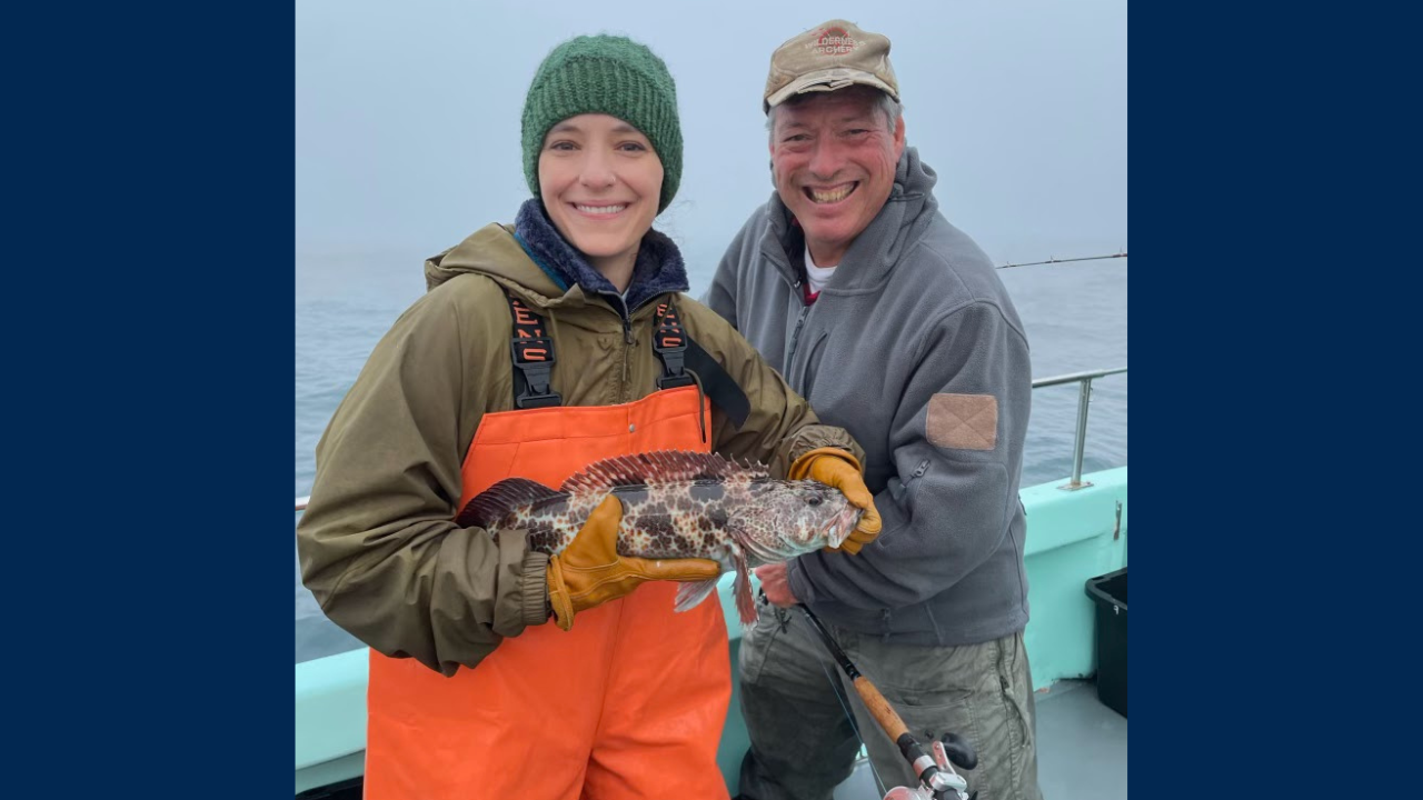 Woman smiling and holding a lingcod next to a man. The man is also smiling and holding a fishing pole.