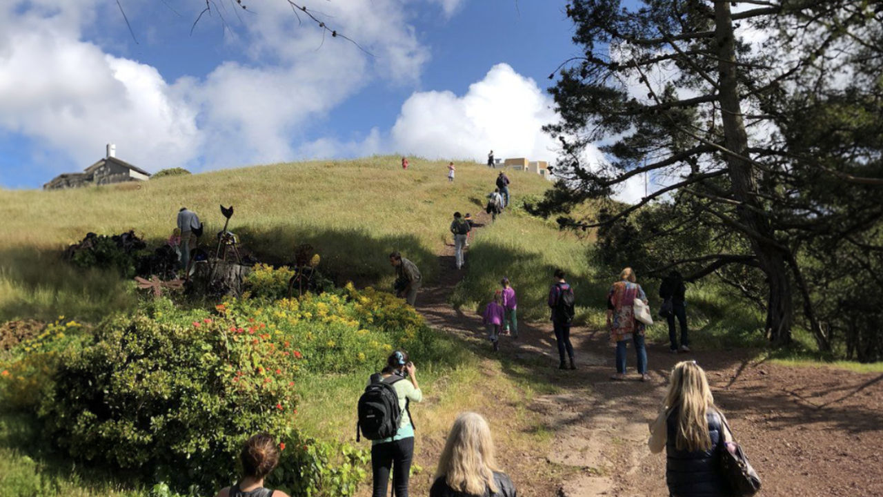 Group of individuals walk on a trail going up a hill. The sky is clear with clouds. 