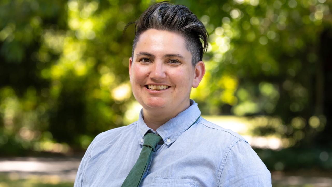 A person with short, dark hair styled upward smiles while standing outdoors. They are wearing a light blue button-up shirt with a green tie. The background shows blurred greenery and natural light.