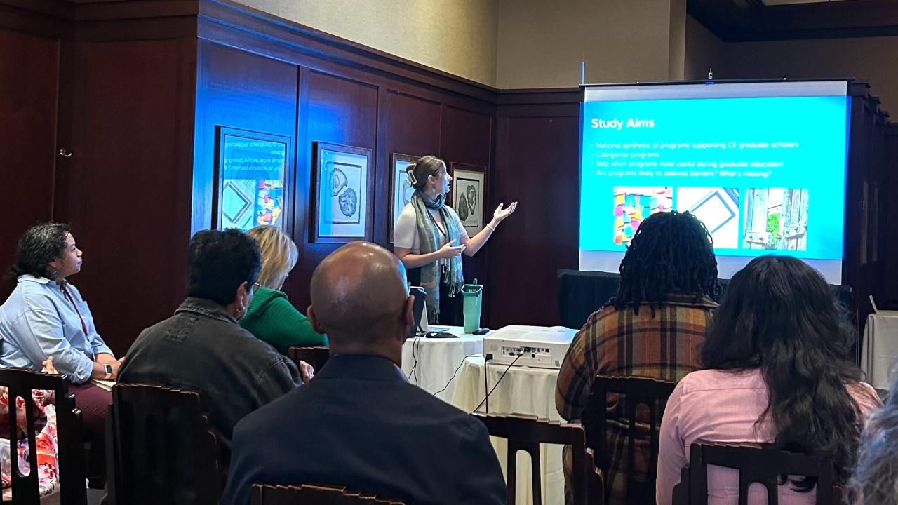 A group of people sits attentively in a conference room, watching a presenter who is standing next to a projection screen. The presenter, wearing a scarf and gesturing with one hand, is discussing a slide titled "Study Aims." The slide includes bullet points and colorful images. Attendees, seated in rows, focus on the presentation, some taking notes. A projector sits on a table in front of the screen, and framed artwork is visible on the wall behind the group.
