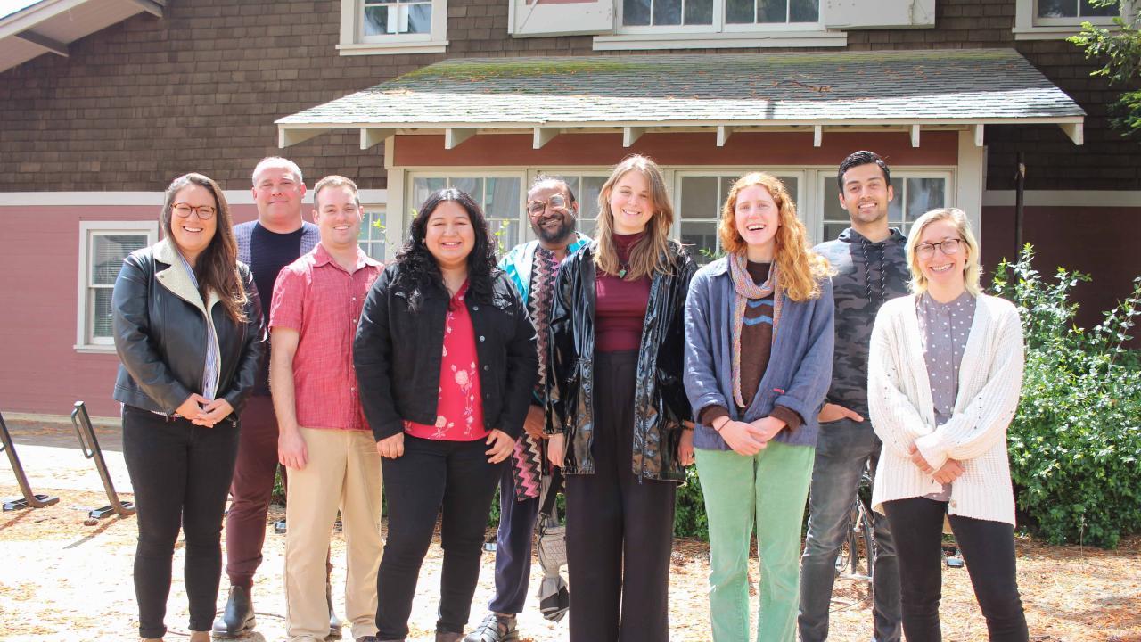 Group of nine individuals smile side by side for a group photo outside of a building