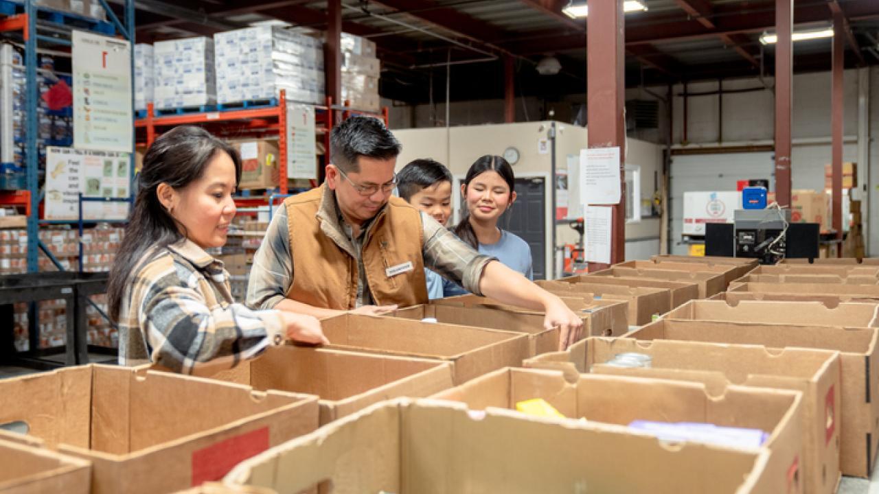 An Asian family of four, consisting of a father, mother and two children, stand at a table full of boxes as they help pack them with non-perishable items at a local Food Bank. They are each dressed casually and smiling at one another as they enjoy their time together helping their community.