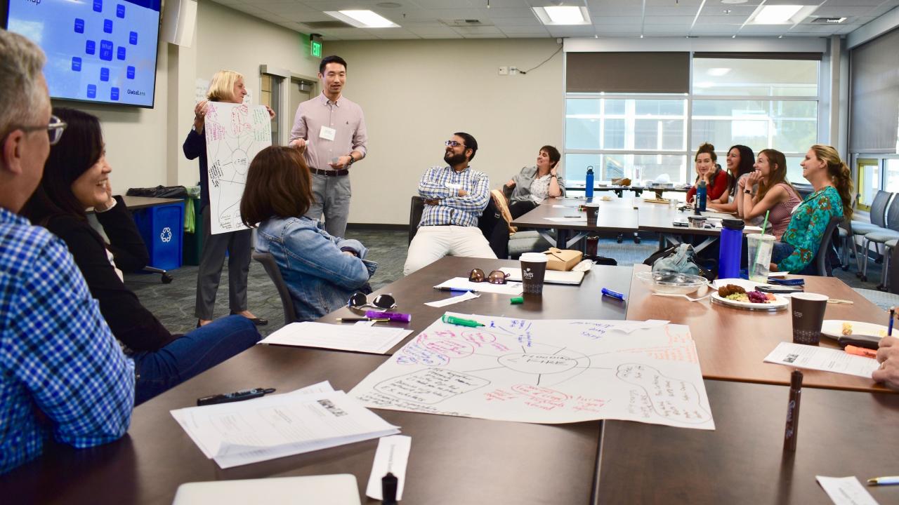 large group of people sitting around a long table looking at a presenter who is holding a poster of a diagram