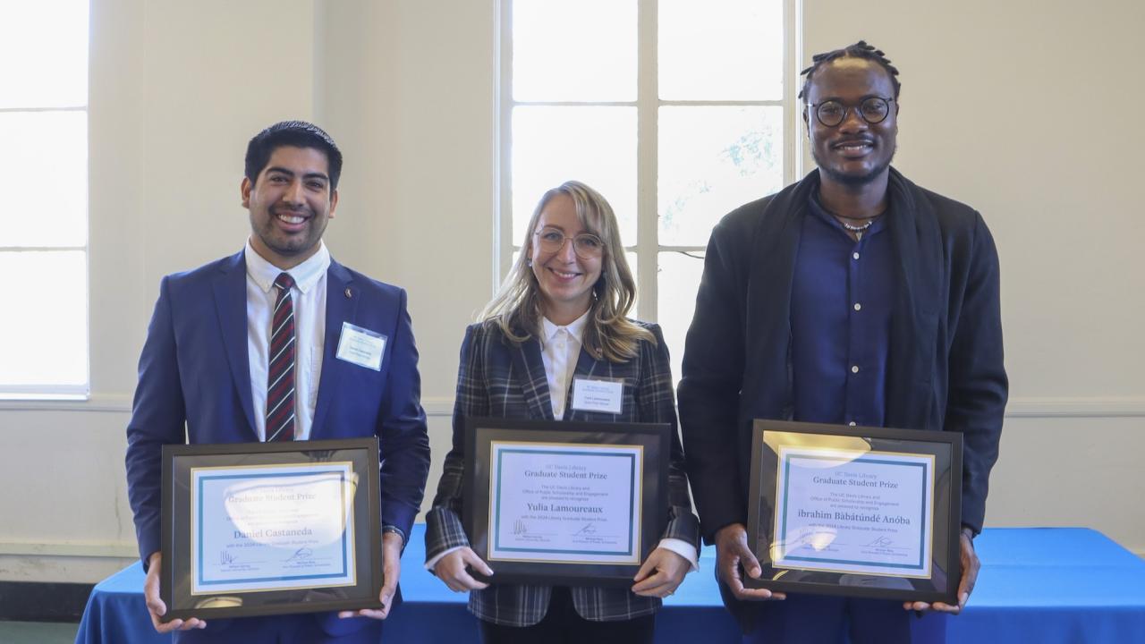 Three individuals stand side by side, each holding a framed certificate. The person on the left wears a blue suit, white shirt, and striped tie. The person in the middle wears glasses, a plaid jacket, and a white blouse. The person on the right wears glasses, a dark jacket, and a blue shirt. They are standing in front of a table with a blue tablecloth in a room with large windows.