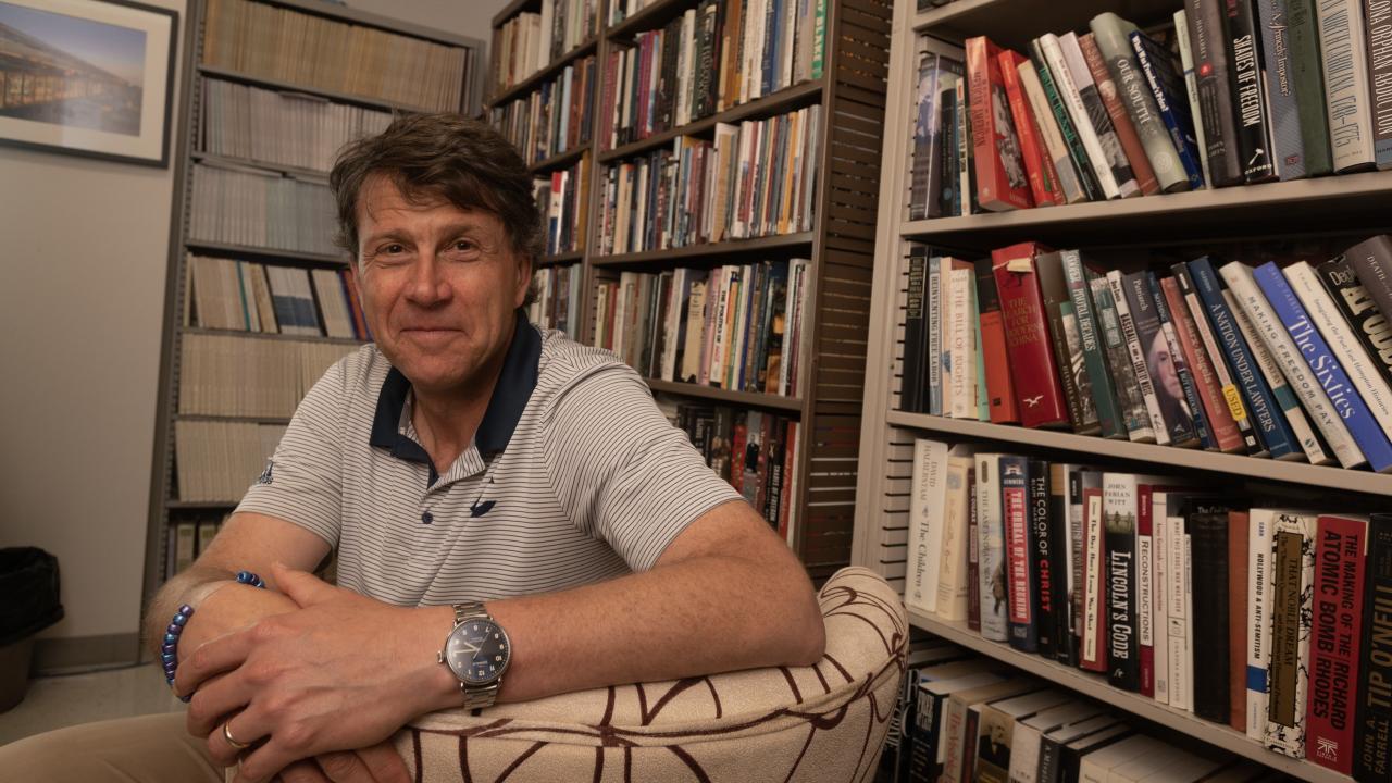 Man in a collared shirt is sitting in a chair. He turns around to smile for a photo while looking directly into the camera. He sits in front of shelves of books.