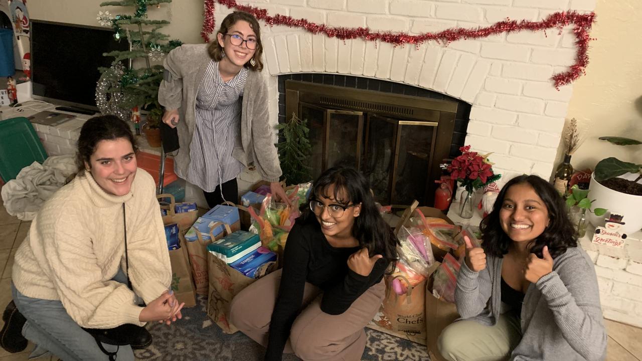 Four young women sit in front of bags of groceries in a decorated holiday living room