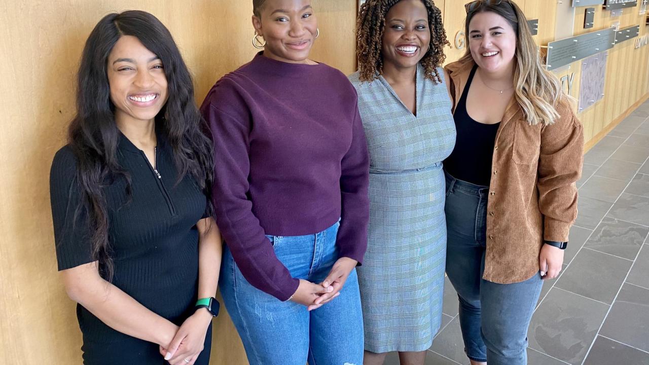 Four women with big smiles stand together for a photo in front of a wall with plaques 