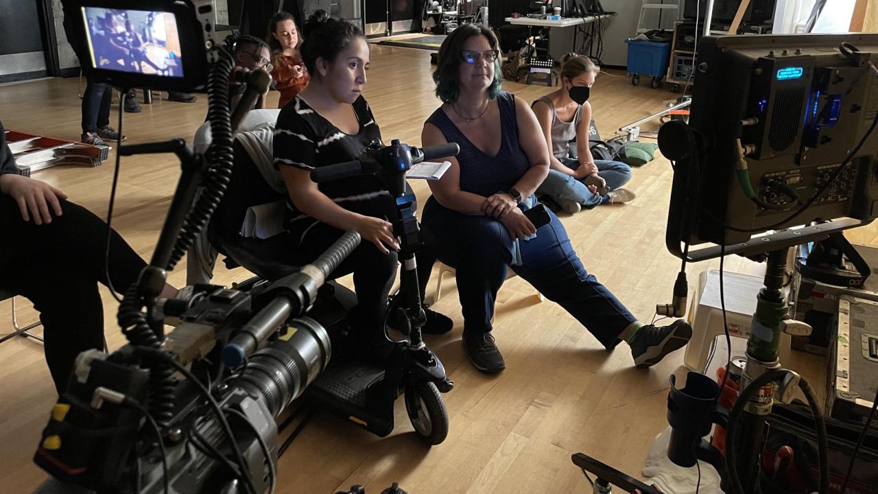 Photo of a set from behind the scenes with two women looking directly at a monitor with other masked individuals in the background