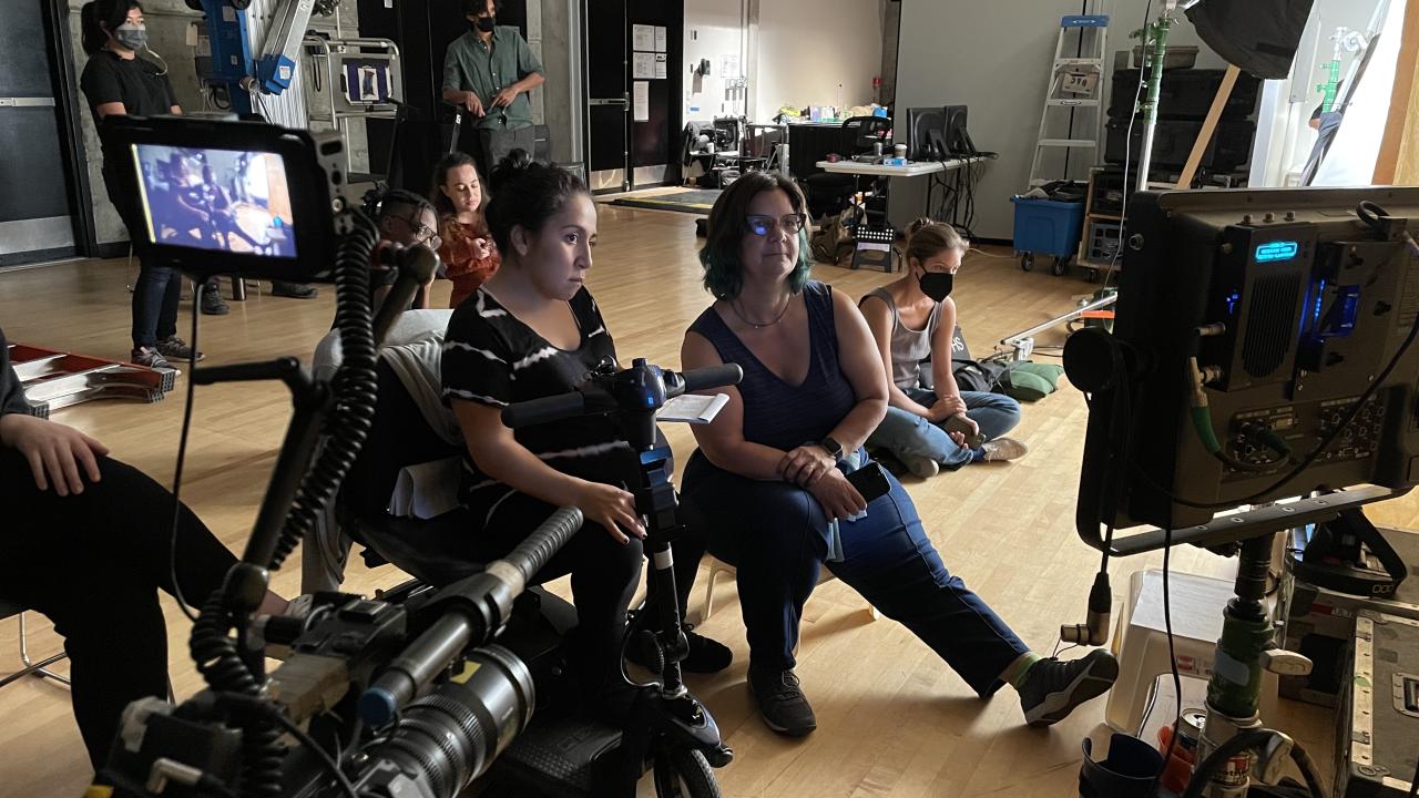 Photo of a set from behind the scenes with two women looking directly at a monitor with other masked individuals in the background