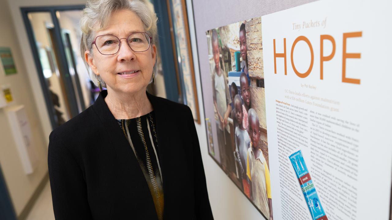 Woman wearing glasses and a blazer smiles softly next to a wall. The wall has a large poster of an article that is titled "Tiny Packets of HOPE"