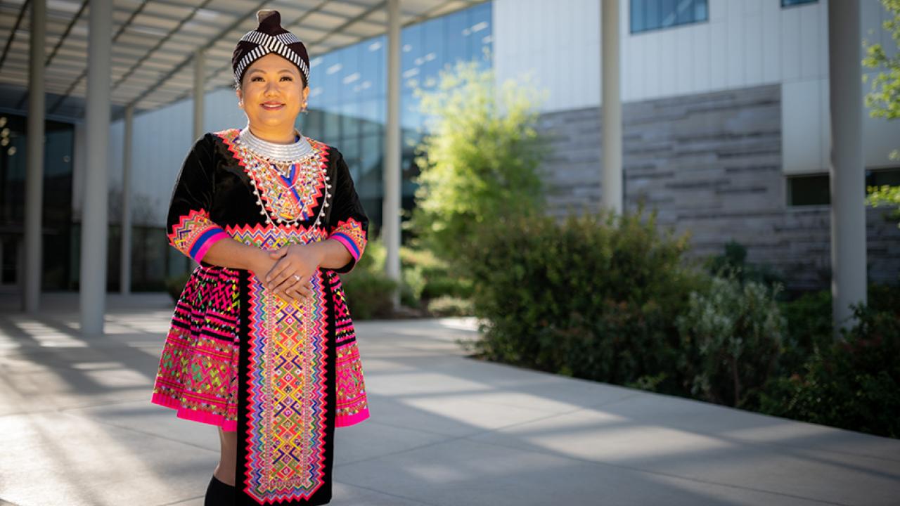 Woman dressed in traditional Hmong clothing smiles outside in front of a building