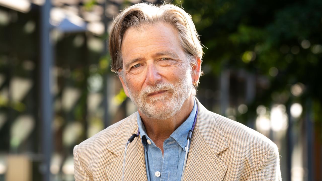 A man with light gray hair and a beard, wearing a beige jacket over a blue shirt, stands outdoors. He has a pair of eyeglasses resting on the front of his jacket. The background shows blurred greenery and parts of a building structure.