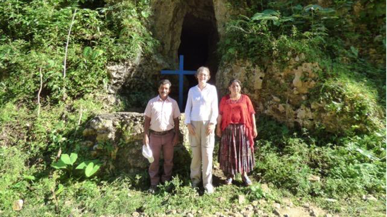 Three individuals stand outside in front of a cave full of leaves and grass